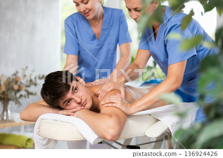Relaxed young man having back massage therapy conducted by two masseuses 136924426