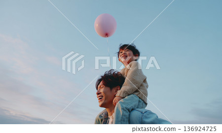 午後，一個男孩騎在父親的肩膀上，手裡拿著一個粉紅色的氣球，在湛藍的天空中大笑。 136924725