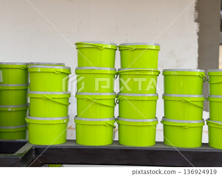 Green plastic buckets stacked in rows against a white wall outdoors. Storage, packaging, and industrial supply represented through containers, order, and retail display. Green plastic buckets stacked in rows against a white wall outdoors. Storage, packaging, and industrial supply represented through containers, order, and retail display. 136924919