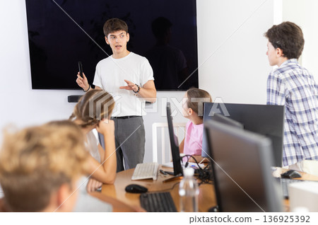 Friendly young teacher standing near an interactive whiteboard telling new material to teenage students in school computer class 136925392