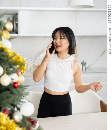 Smiling young Asian girl speaking on the phone standing by Xmas tree 136925393