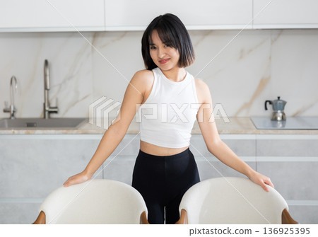 Smiling young Asian girl standing at table in the kitchen Smiling young Asian girl standing at table in the kitchen 136925395