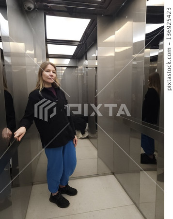 A woman stands inside a modern elevator with metallic mirrored walls, urban lifestyle, daily commute, modern interior. A woman stands inside a modern elevator with metallic mirrored walls, urban lifestyle, daily commute, modern interior. 136925423