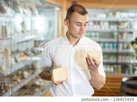 Attentive young man purchaser choosing cheese in large supermarket Attentive young man purchaser choosing cheese in large supermarket 136925429