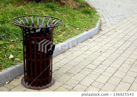 Trash Can Placed Near an Alley in a Park Surrounded by Grass and Pavement 136925543
