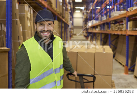 Warehouse worker with pallet trolley moving boxes between shelves in storage 136926298