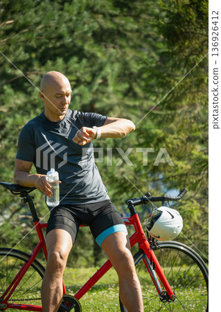 Mature cyclist resting on bicycle checking smartwatch and holding water bottle Mature cyclist resting on bicycle checking smartwatch and holding water bottle 136926412