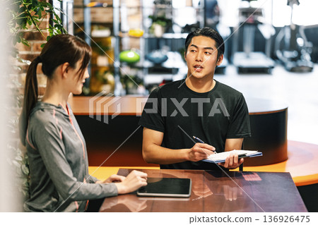 A male trainer giving an explanation at the reception desk of a sports gym A male trainer giving an explanation at the reception desk of a sports gym 136926475