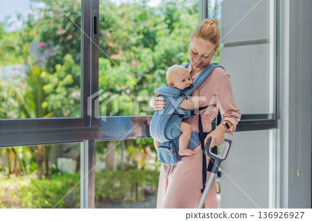 Woman vacuuming a room while carrying her baby in an ergonomic carrier. Modern motherhood, multitasking parent, home chores with infant, balance of childcare and housework concept. Woman vacuuming a room while carrying her baby in an ergonomic carrier. Modern motherhood, multitasking parent, home chores with infant, balance of childcare and housework concept. 136926927
