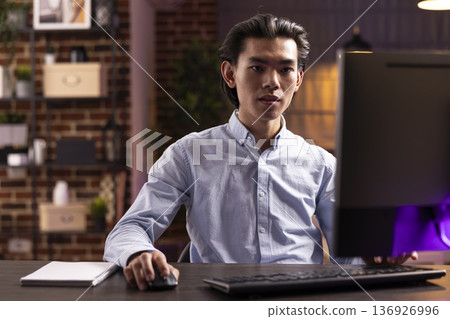 Focused businessman works on digital tasks, sitting at modern desk in home office. Handsome asian entrepreneur reviews marketing data and emails to improve small business strategies. 136926996