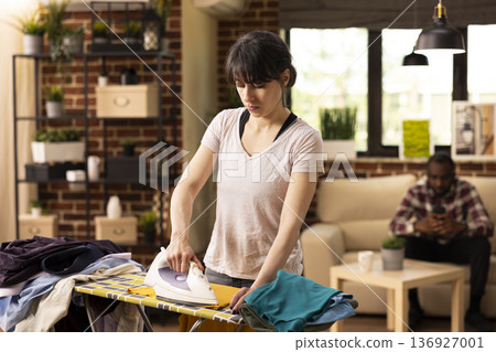 White woman ironing shirt on board, using electric steam iron to remove wrinkles after washing laundry. Beautiful lady focused on household chores, pressing clothes neatly at home in modern apartment. White woman ironing shirt on board, using electric steam iron to remove wrinkles after washing laundry. Beautiful lady focused on household chores, pressing clothes neatly at home in modern apartment. 136927001