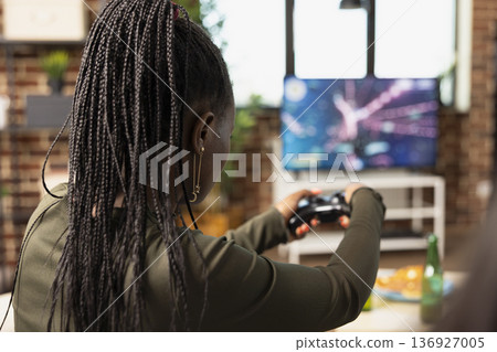 Young black woman plays console game on couch, holding joystick and enjoying fun digital action. Diverse friends sits with wireless controllers, competing in online video game during casual hangout. Young black woman plays console game on couch, holding joystick and enjoying fun digital action. Diverse friends sits with wireless controllers, competing in online video game during casual hangout. 136927005