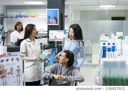 Male wheelchair user receives medication guidance from pharmacist while accompanied by his mother. Pharmacist assists both caregiver and patient in choosing the most suitable medication for treatment. Male wheelchair user receives medication guidance from pharmacist while accompanied by his mother. Pharmacist assists both caregiver and patient in choosing the most suitable medication for treatment. 136927012