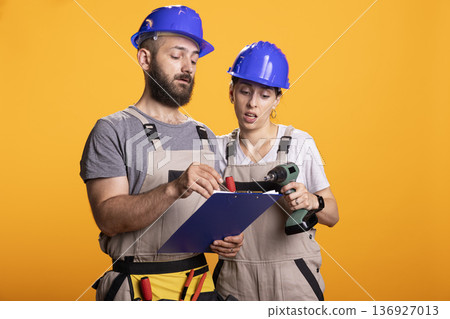 Male and female builders with clipboard, discussing upcoming tasks against isolated background. Caucasian coworkers in protective gear show teamwork and shared responsibility in construction planning. Male and female builders with clipboard, discussing upcoming tasks against isolated background. Caucasian coworkers in protective gear show teamwork and shared responsibility in construction planning. 136927013