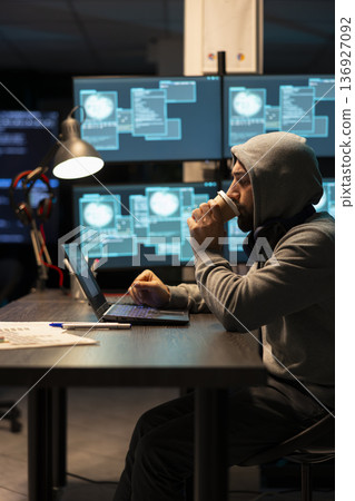 Male programmer works late in office, sipping coffee while reviewing coding software on laptop. Caucasian man wearing hoodie, drinks caffeine and monitors encrypted data on digital device. Male programmer works late in office, sipping coffee while reviewing coding software on laptop. Caucasian man wearing hoodie, drinks caffeine and monitors encrypted data on digital device. 136927092