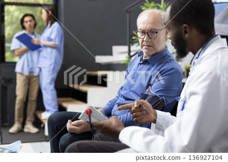 Experienced doctor in lab coat measures blood pressure of caucasian senior man during routine medical checkup. The scene reflects professional healthcare, preventive care and trust in modern clinic. Experienced doctor in lab coat measures blood pressure of caucasian senior man during routine medical checkup. The scene reflects professional healthcare, preventive care and trust in modern clinic. 136927104