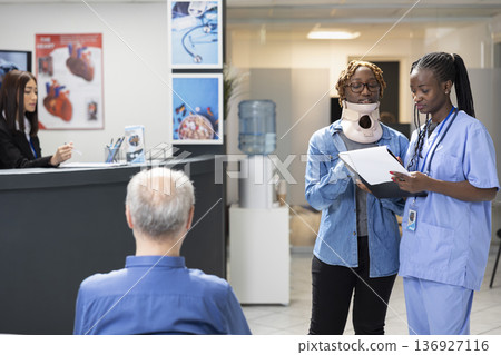 Healthcare worker discussing medical care information with black woman wearing cervical collar in hospital reception. Injured female patient confirming doctors appointment with nurse holding clipboard Healthcare worker discussing medical care information with black woman wearing cervical collar in hospital reception. Injured female patient confirming doctors appointment with nurse holding clipboard 136927116