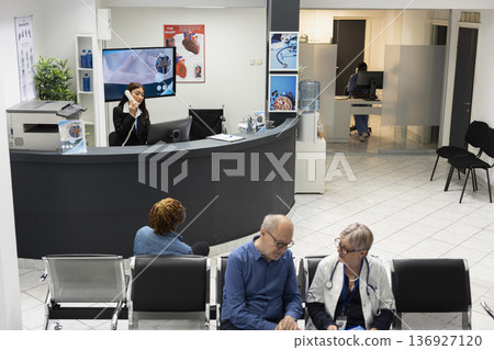 Diverse medical staff and patients in clinic lobby during checkup visits as receptionist answers phone call. Senior doctor discusses diagnosis with elderly man as black woman waits for consultation. Diverse medical staff and patients in clinic lobby during checkup visits as receptionist answers phone call. Senior doctor discusses diagnosis with elderly man as black woman waits for consultation. 136927120
