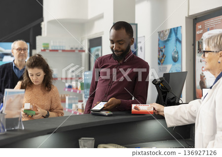 African american man pays for pharmacy items using smartphone, demonstrating cashless payment methods. Senior female pharmacist provides efficient customer service as black customer purchases medicine African american man pays for pharmacy items using smartphone, demonstrating cashless payment methods. Senior female pharmacist provides efficient customer service as black customer purchases medicine 136927126