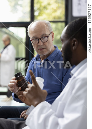 Professional physician discusses medical treatment details with male patient in hospital lobby. Focused consultation includes pill bottle explanation, dosage guidance and supportive healthcare advice. Professional physician discusses medical treatment details with male patient in hospital lobby. Focused consultation includes pill bottle explanation, dosage guidance and supportive healthcare advice. 136927127