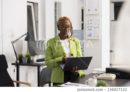 Marketing specialist stands focused on reviewing company analytics and supporting team with strategic decisions. Female manager maintaining professional posture with laptop in hand, checking reports. Marketing specialist stands focused on reviewing company analytics and supporting team with strategic decisions. Female manager maintaining professional posture with laptop in hand, checking reports. 136927132