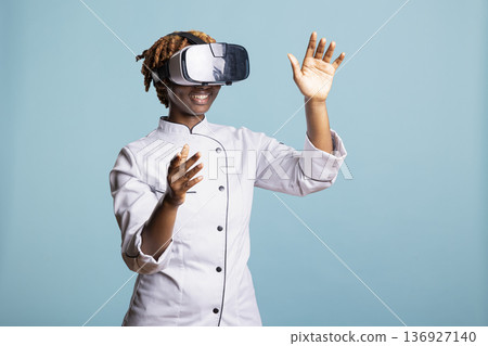 Black woman chef interacts with virtual reality interface, exploring digital cooking techniques. Female cook uses AR technology to practice culinary skills and prepare for upcoming competitions. Black woman chef interacts with virtual reality interface, exploring digital cooking techniques. Female cook uses AR technology to practice culinary skills and prepare for upcoming competitions. 136927140