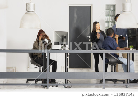 Black woman appears overwhelmed while working in diverse workspace, struggling to complete programming projects. Female developer pauses to ponder debugging coding scripts to achieve a functional app. Black woman appears overwhelmed while working in diverse workspace, struggling to complete programming projects. Female developer pauses to ponder debugging coding scripts to achieve a functional app. 136927142