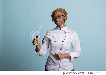 Black professional cook poses confidently with kitchen knife in hand, standing tall in studio. African american female chef expressing focus and readiness, reflecting confidence in culinary industry. 136927143