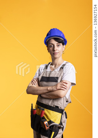 White woman dressed in overalls and safety helmet stands confidently with arms crossed in studio. Skilled contractor in workwear poses for advertising, representing renovation and handyman services White woman dressed in overalls and safety helmet stands confidently with arms crossed in studio. Skilled contractor in workwear poses for advertising, representing renovation and handyman services 136927154