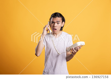 Portrait of asian man holding telephone and reacting in disbelief. Male model shows confusion and surprise after hearing shocking or unpleasant news, representing communication and emotional response. Portrait of asian man holding telephone and reacting in disbelief. Male model shows confusion and surprise after hearing shocking or unpleasant news, representing communication and emotional response. 136927158