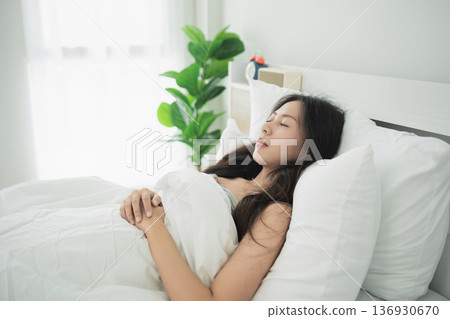Young woman peacefully sleeping in a cozy bedroom with bright natural light, showcasing tranquility and comfort in a modern home setting. 136930670