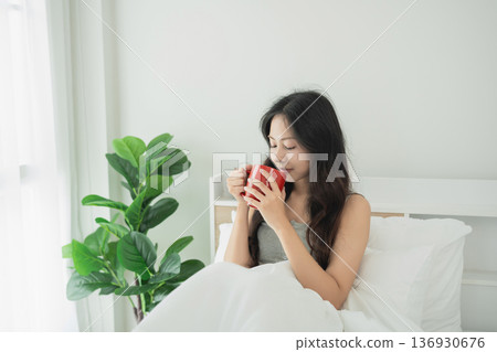 Young woman enjoying a warm beverage in a bright, cozy bedroom with white bedding and green plant in natural light for relaxation and comfort Young woman enjoying a warm beverage in a bright, cozy bedroom with white bedding and green plant in natural light for relaxation and comfort 136930676