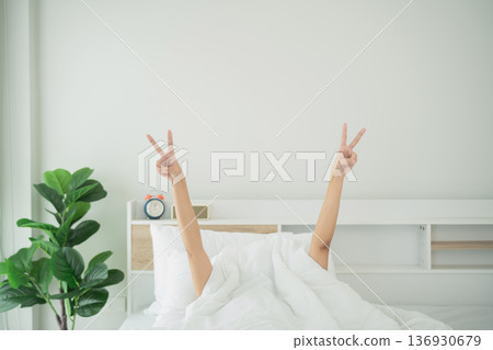 Young person joyfully relaxing in a cozy bed, morning light illuminating fresh white linens and fresh indoor plant, offering a peaceful and calm atmosphere 136930679