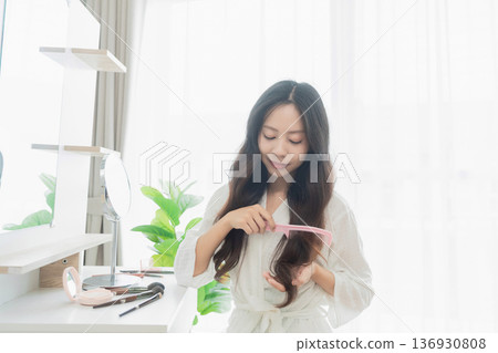 Young woman in light robe combing her long hair in a bright and airy room with natural light and greenery decorating the space Young woman in light robe combing her long hair in a bright and airy room with natural light and greenery decorating the space 136930808
