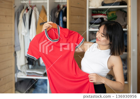 Happy young Asian girl trying to choose a dress standing by wardrobe Happy young Asian girl trying to choose a dress standing by wardrobe 136930956