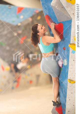 Woman in comfortable athletic clothing moving carefully on bouldering wall clinging to colorful hand and foot holds Woman in comfortable athletic clothing moving carefully on bouldering wall clinging to colorful hand and foot holds 136931037