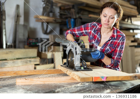 Working at sawmill - woman saws wooden board using circular saw 136931186