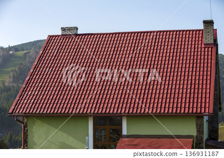 Detail of new modern house cottage corner with stucco walls decorated with natural stones, red shingled roof and rain gutter pipe system on blurred spruce forest copy space background. Detail of new modern house cottage corner with stucco walls decorated with natural stones, red shingled roof and rain gutter pipe system on blurred spruce forest copy space background. 136931187