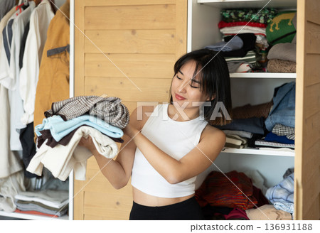 Happy young Asian girl holding some items of clothing by the wardrobe 136931188
