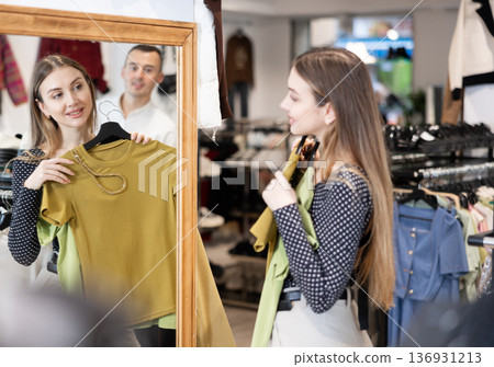Girl stands in front of mirror hold hanger with blouse, puts it to herself and admires her reflection. 136931213