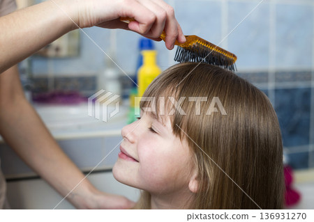 Mother hand with brush combing long fair hair of cute child girl after bath on blurred interior background. Mother hand with brush combing long fair hair of cute child girl after bath on blurred interior background. 136931270