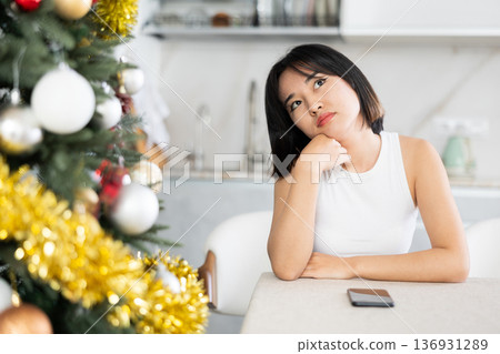 Sad young Asian girl waiting for phone call sitting at table by Xmas tree 136931289
