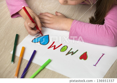View from above of childs hands drawing with colorful crayons I love Mom on white paper. Art education, creativity concept. 136931337