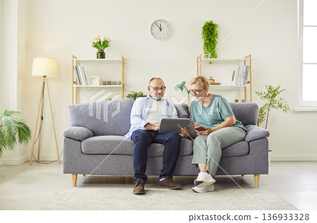 Senior couple sitting on couch at home using tablet and smartphone together Senior couple sitting on couch at home using tablet and smartphone together 136933328