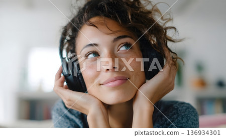 A thoughtful young woman with curly hair and freckles listens intently with black over-ear headphones, gazing upward while enjoying music or a peaceful podcast. AI Generated A thoughtful young woman with curly hair and freckles listens intently with black over-ear headphones, gazing upward while enjoying music or a peaceful podcast. AI Generated 136934301