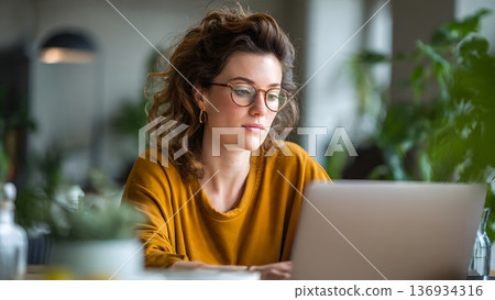 A young woman with curly hair and stylish glasses intently uses a laptop in a bright indoor setting, displaying deep concentration on her professional work. AI Generated 136934316