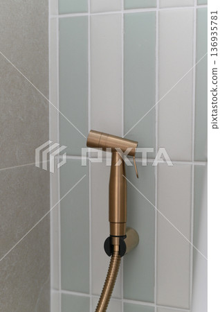 Brass handheld bidet sprayer mounted on bathroom wall, modern sanitary fixture with flexible hose, vertical white and light green tiles, clean and minimalistic design 136935781