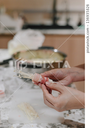 Preparing enoki mushroom bacon roll, hands wrapping fresh enoki mushroom with sliced bacon on kitchen counter, stainless steel pot and cutting board in background, home cooking concept Preparing enoki mushroom bacon roll, hands wrapping fresh enoki mushroom with sliced bacon on kitchen counter, stainless steel pot and cutting board in background, home cooking concept 136935826