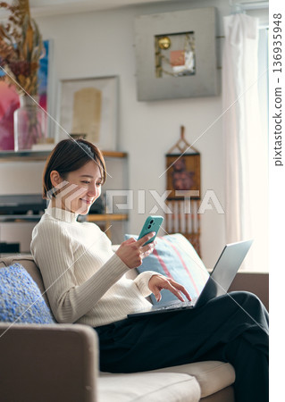 一名女子一邊操作電腦一邊拿著智慧型手機。 一名女子一邊操作電腦一邊拿著智慧型手機。 136935948