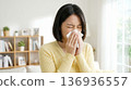 A Japanese woman wearing a yellow cardigan blowing her nose with a tissue in the living room. Image of hay fever or a cold. 136936557
